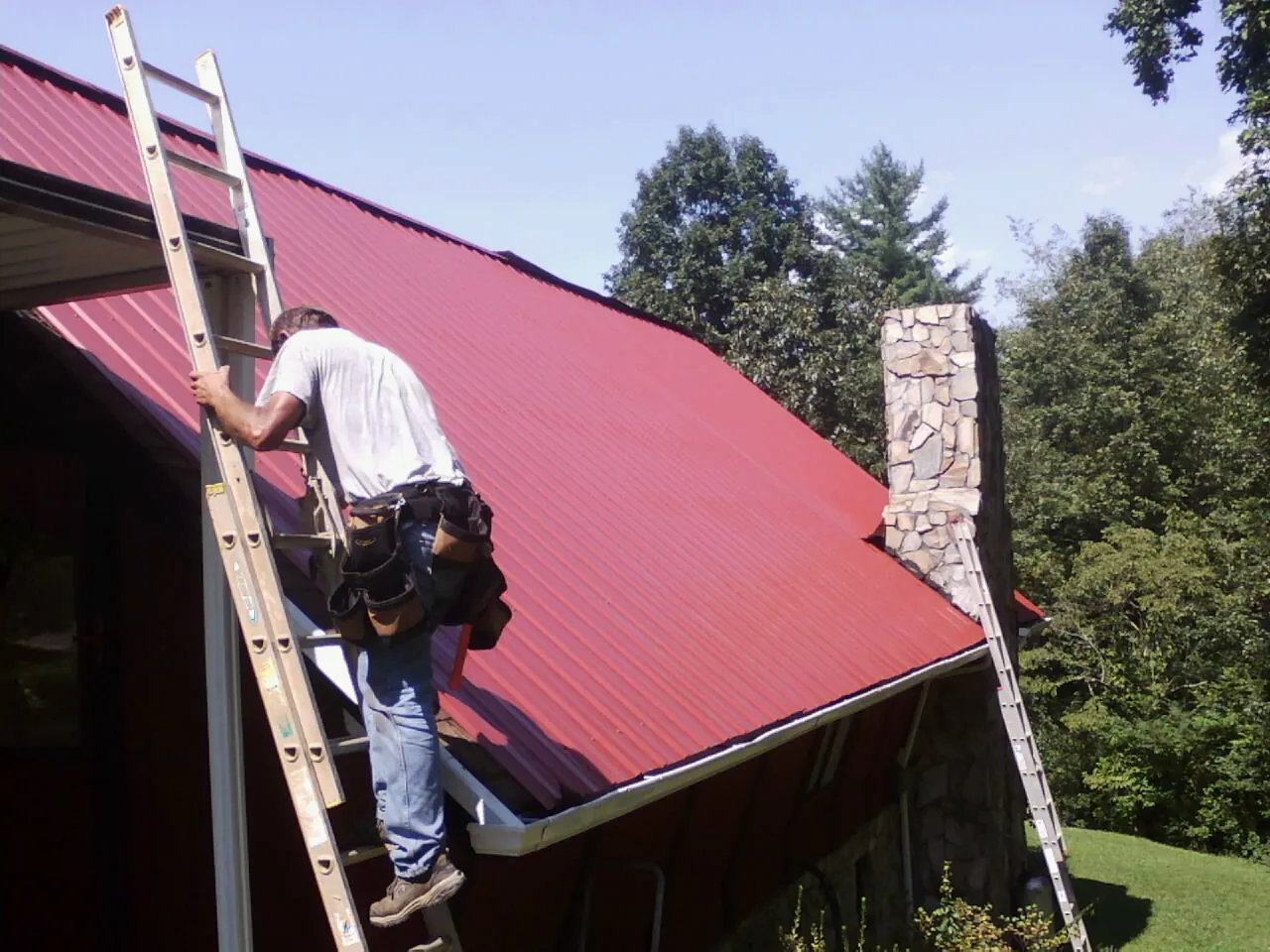 Roofer on a ladder beside a red metal roof and stone chimney — Soffit & Fascia Repair by Roofing Brothers in Morral